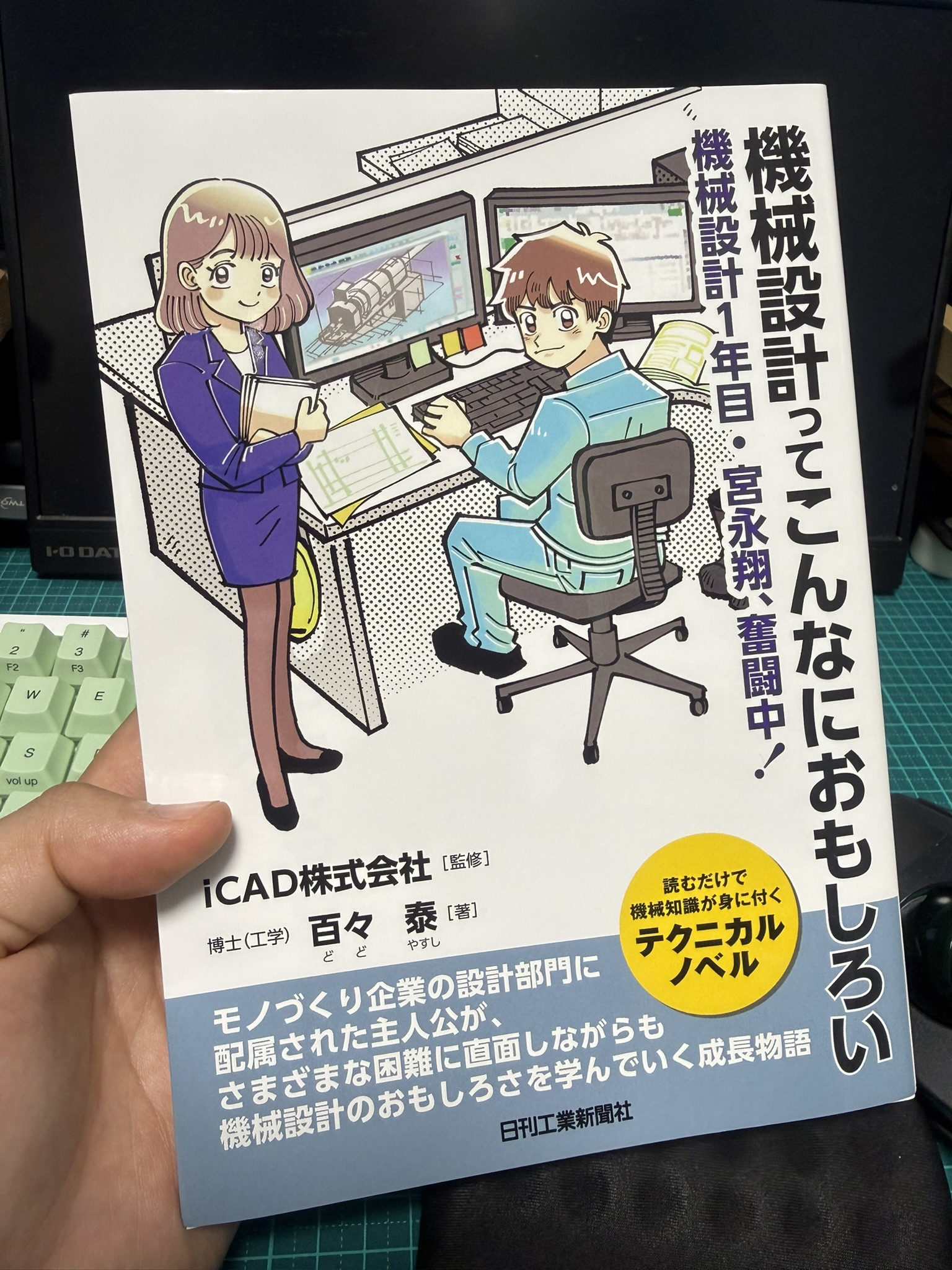 書籍『機械設計ってこんなにおもしろい―機械設計1年目・宮永翔、奮闘中!』の表紙を手に持った写真。iCAD株式会社監修、百々泰著、日刊工業新聞社のテクニカルノベル